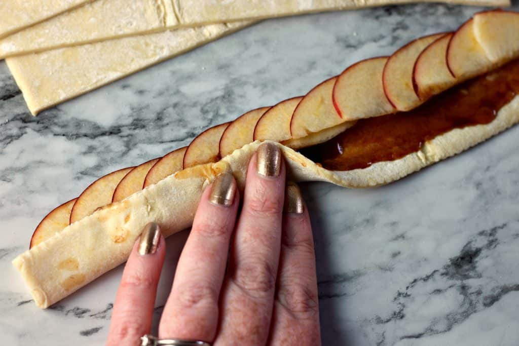 Folding puff pastry over thinly sliced apples on a marble counter